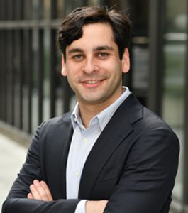 Man in suit jacket and shirt with no tie with arms crossed standing in front of a fence.