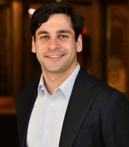 Man in jacket and shirt with no tie in front of a dark background
