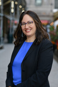 Woman in black suit and blue shirt standing on a street.